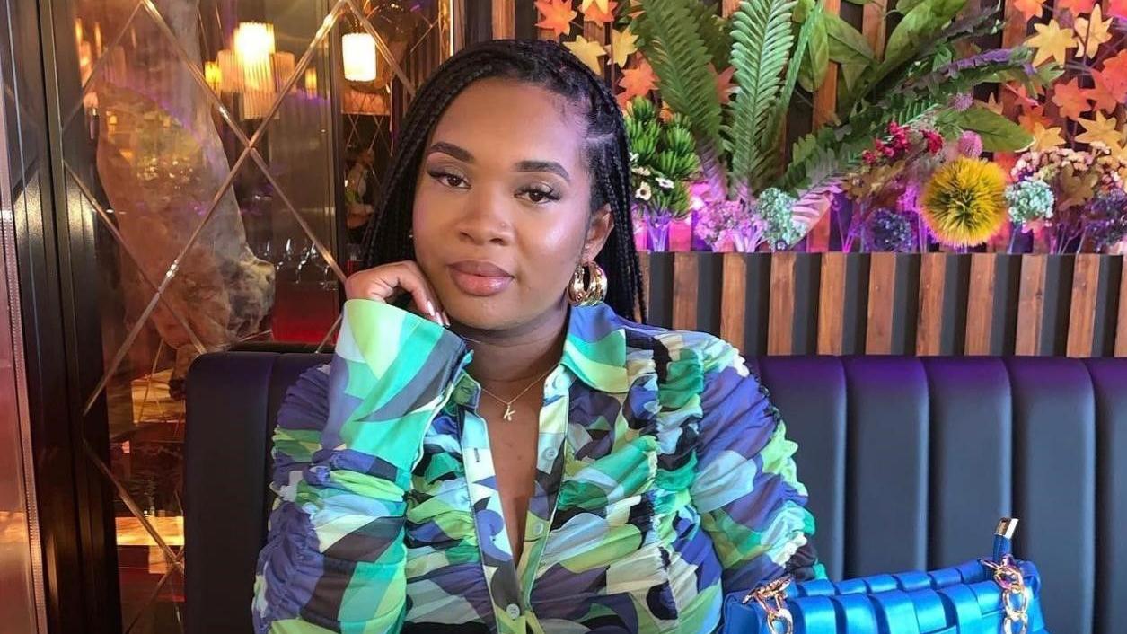 A woman with braided hair sits at a restaurant table. She is wearing a colorful patterned blouse in shades of blue, green, and purple, along with gold hoop earrings and a delicate necklace with a small pendant. A bright blue handbag rests on the seat beside her. Behind her is a wall decorated with plants and flowers, illuminated by soft lighting.