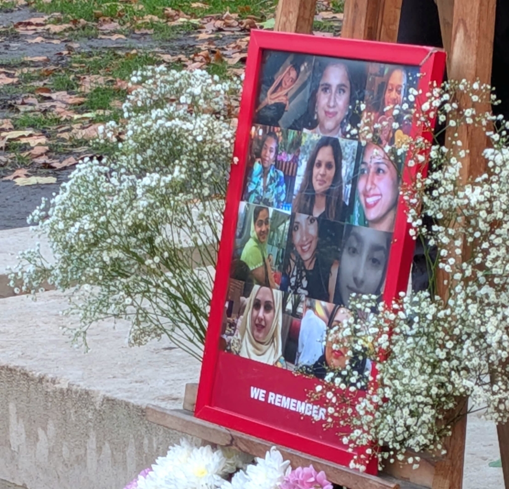 A memorial display with a red-framed collage of women’s faces on a wooden stand, surrounded by white baby’s breath and colorful bouquets of flowers.